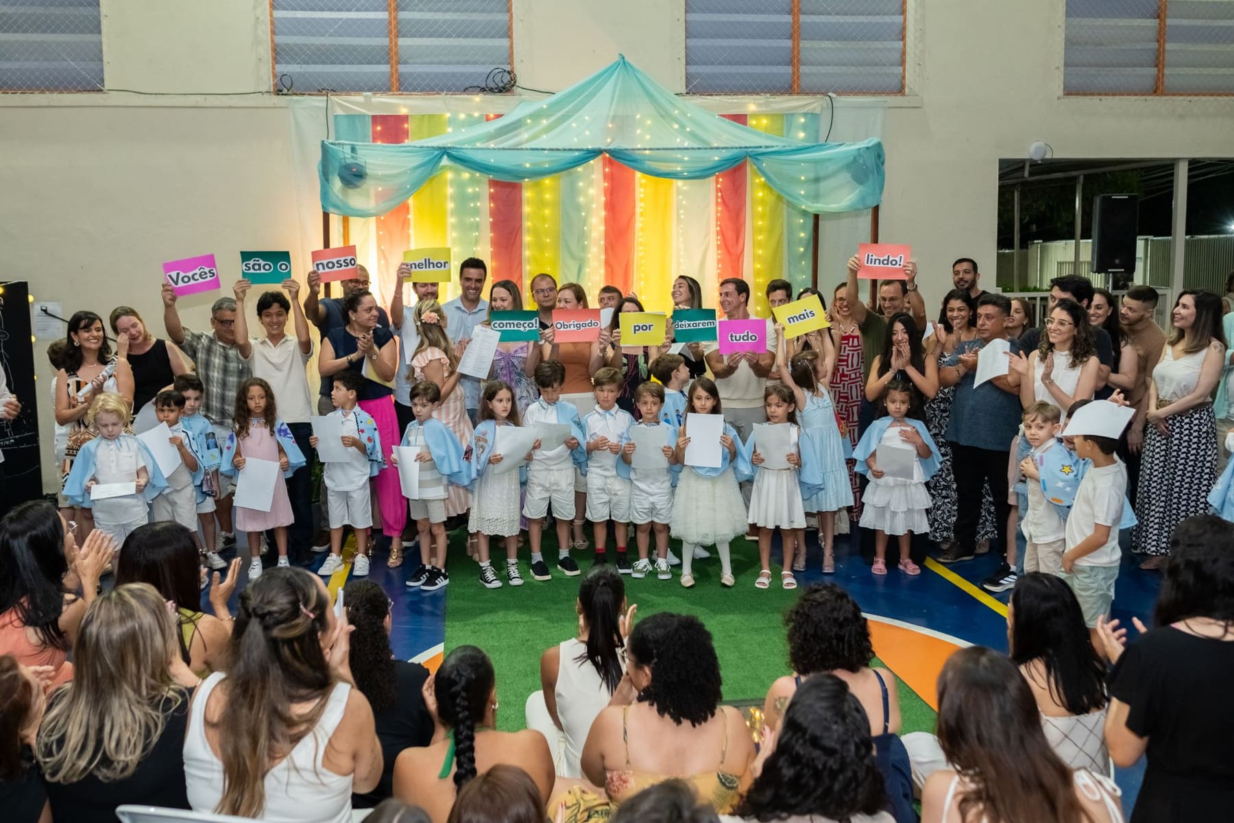 Turma da formatura no palco, com familiares segurando plaquinhas com a frase: vocês são o nosso melhor começo